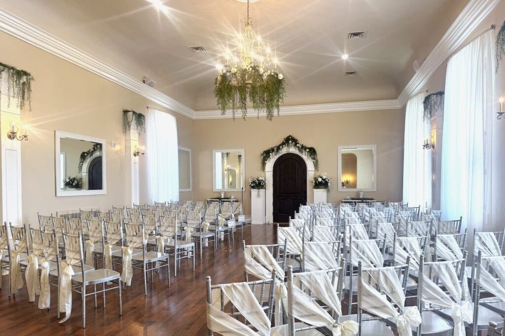 A brightly-lit ceremony space is set up with silver chairs and greenery