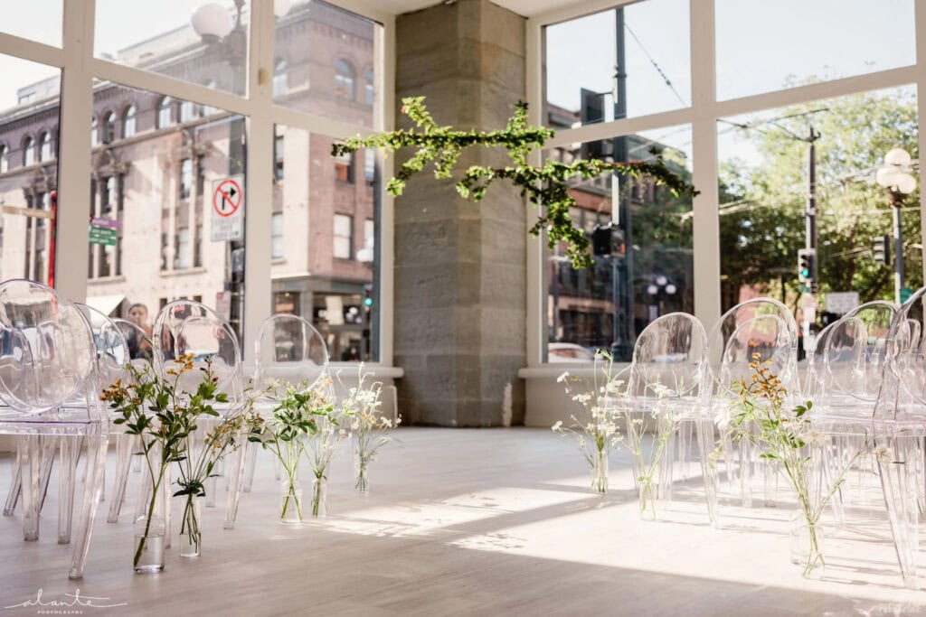 Acrylic ghost chairs line a simply-decorated aisle at a brightly-lit ceremony venue in the city