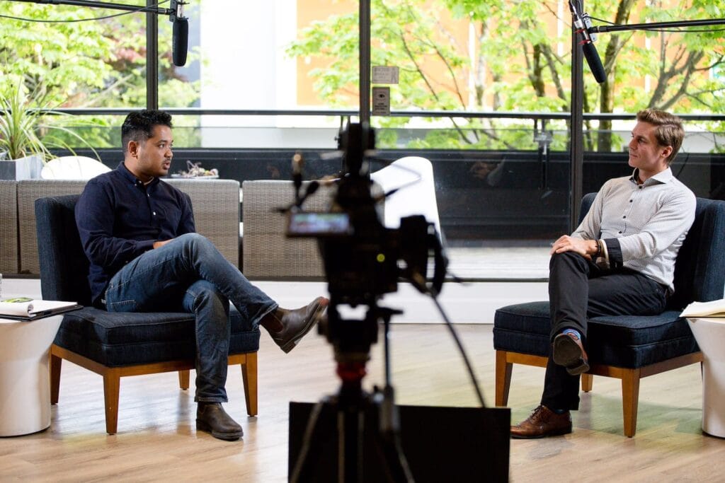 To men sit across from each other while having a conversation while being recorded on camera in a sunlit commercial space