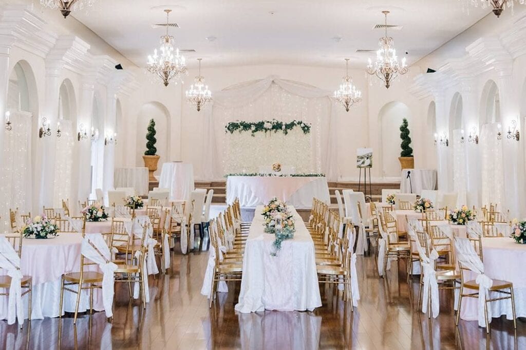 An all-white venue space is decorated with gold chairs, white and pink linens and pale-colored flowers