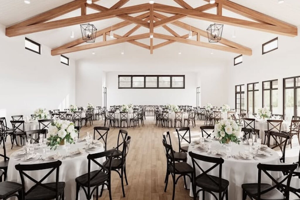 Wooden beams highlight a bright white venue space set up with numerous round tables, each surrounded by black wooden chairs