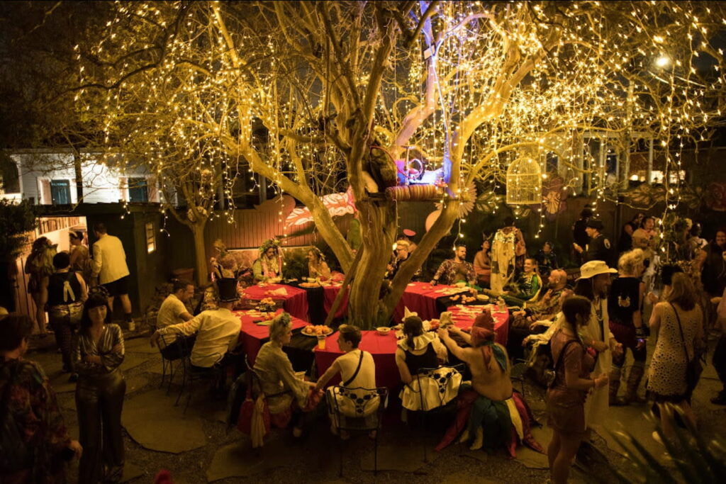 People gather at a round table encircling a giant tree draped in lights at an outdoor event in New Orleans