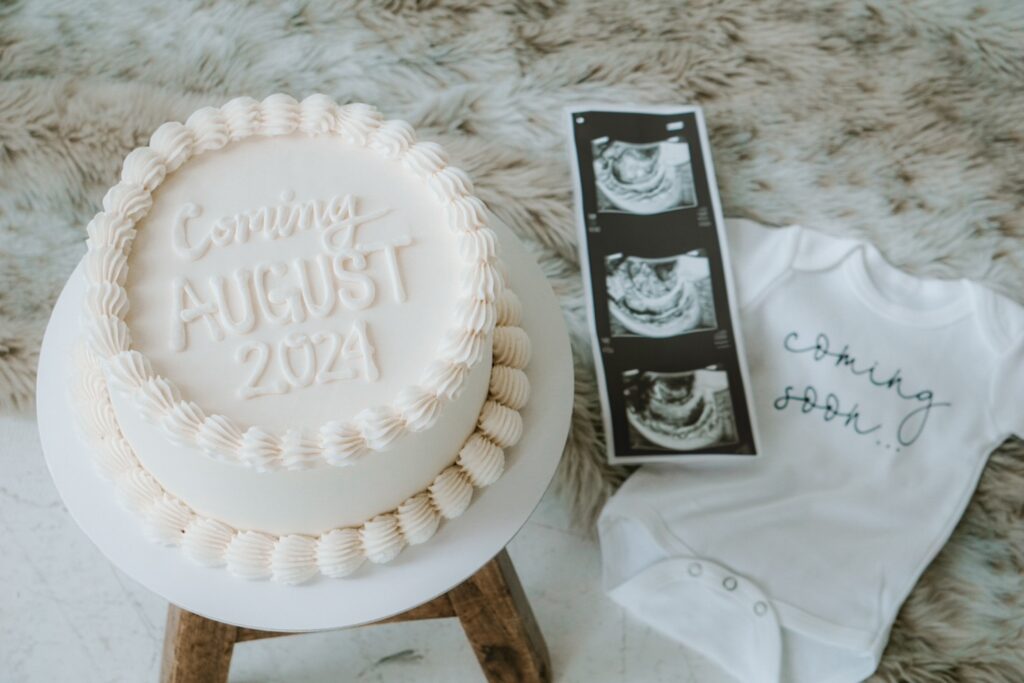 A white cake reading "Coming August 2024" sits next to a white onesie that says "Coming Soon" and an ultrasound picture