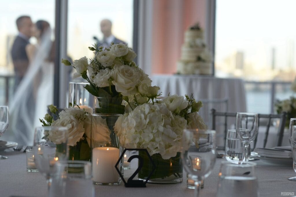 White floral centerpieces are surrounded by votive candles on a table. In the background, the wedding cake and bride can be seen