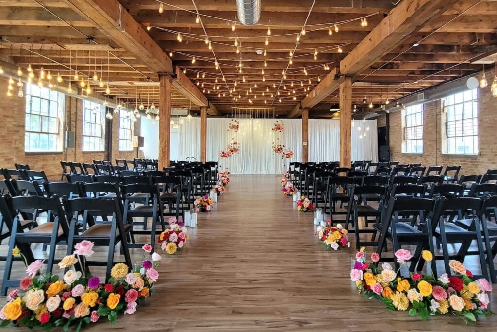 A wedding ceremony is set up with black wooden chairs, brightly-colored flowers and string lights in a warehouse