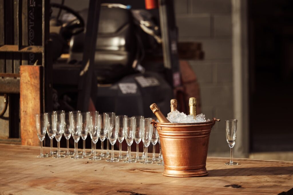 A bucket of champagne sits atop a wooden bar next to a row of champagne glasses