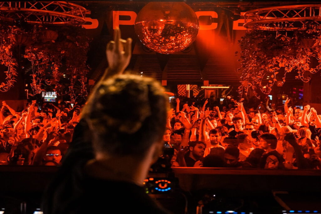 A crowd dances to a DJ, whose silhouette can be seen in the foreground