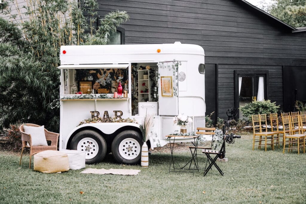 A small bar cart is set up in front of a dark building by gold ceremony chairs surrounded by a bistro table and other seating areas