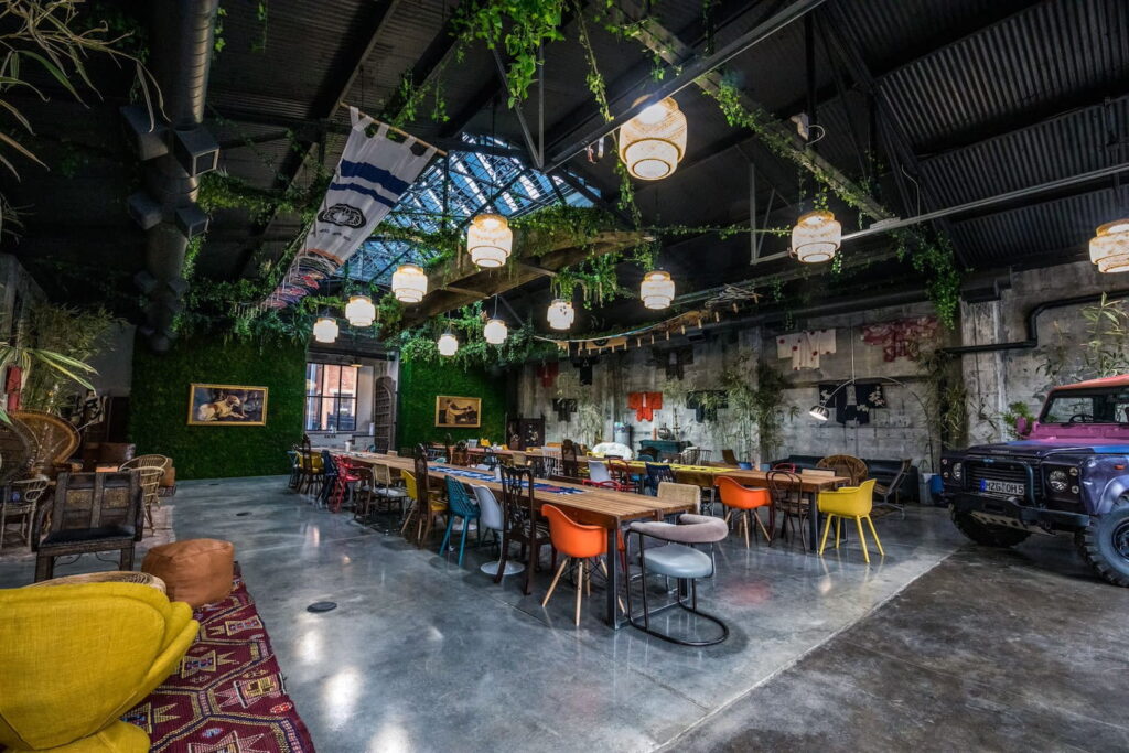 A converted warehouse is set up for an event with long tables, colorful chairs and greenery on walls and the ceiling