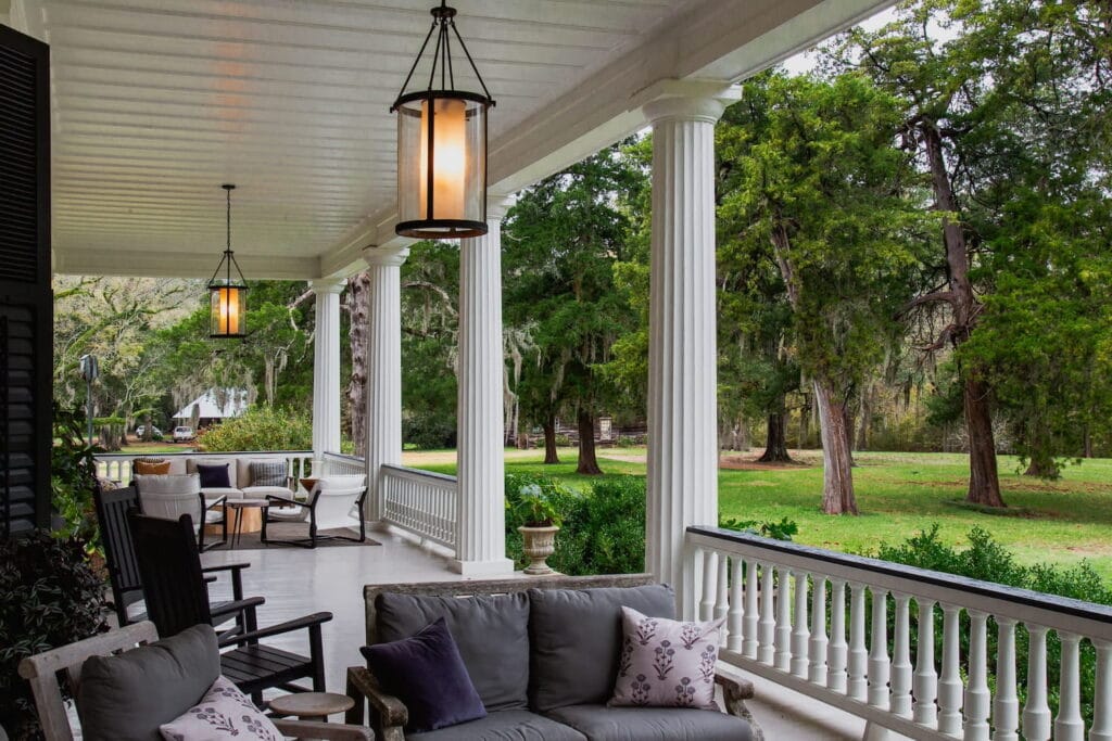 A large veranda at a southern estate with rocking chairs and different seating areas looking out onto a lawn covered in large trees
