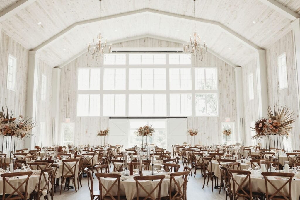 Bright, white barn with a huge wall of windows decorated for a wedding reception with wooden chairs and boho-chic centerpieces