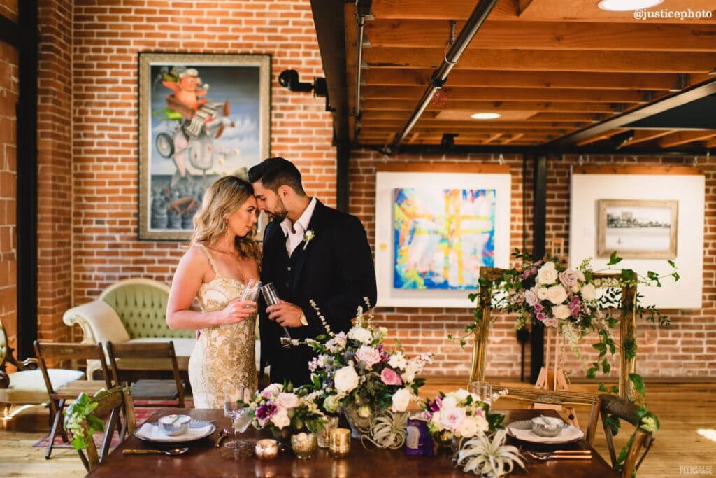 A couple steal a private moment standing at a reception table decorated in dramatic florals at an art gallery venue with brick walls