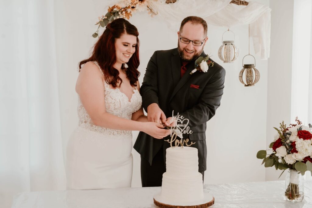 A bride and groom stand smiling as they cut their wedding cake