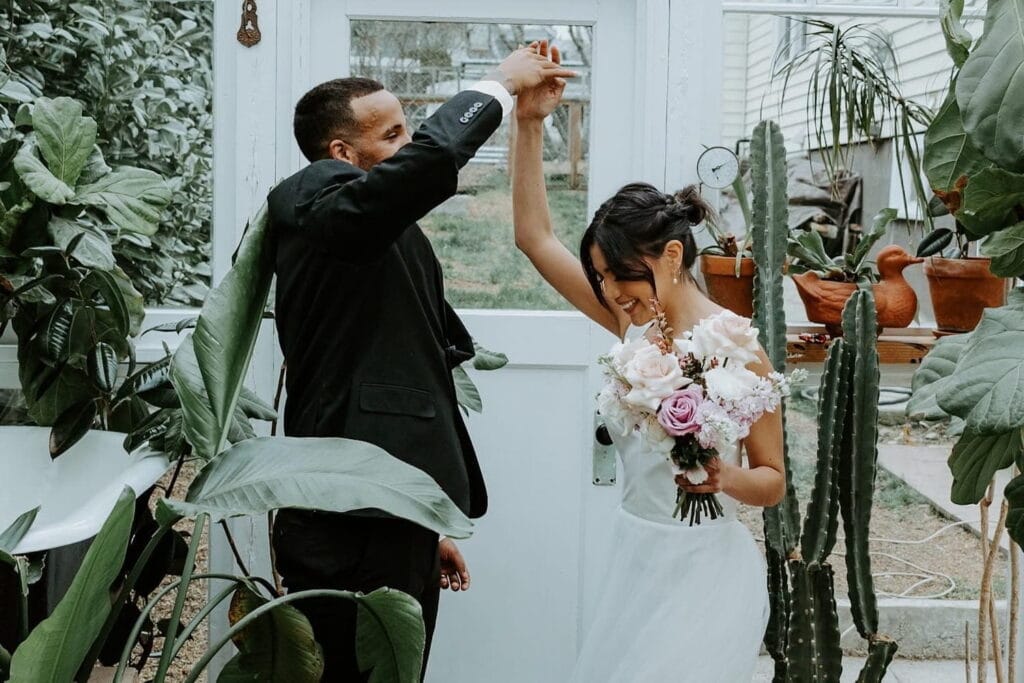 A groom spins his wife inside a greenhouse full of lush greenery
