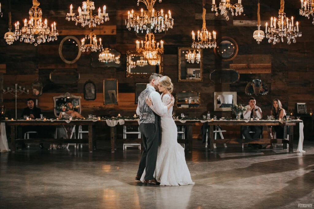 A bride and groom slow dance in front of a long reception table in a romantic venue decorated with barn wood, chandeliers and mirrors