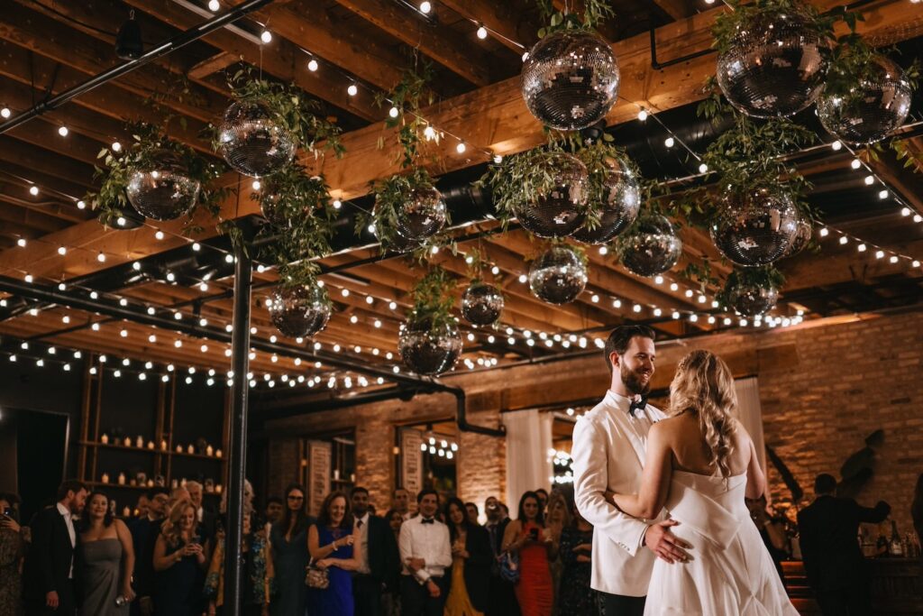 A newlywed couple dances in front of friends and family under a ceiling of string lights and disco balls