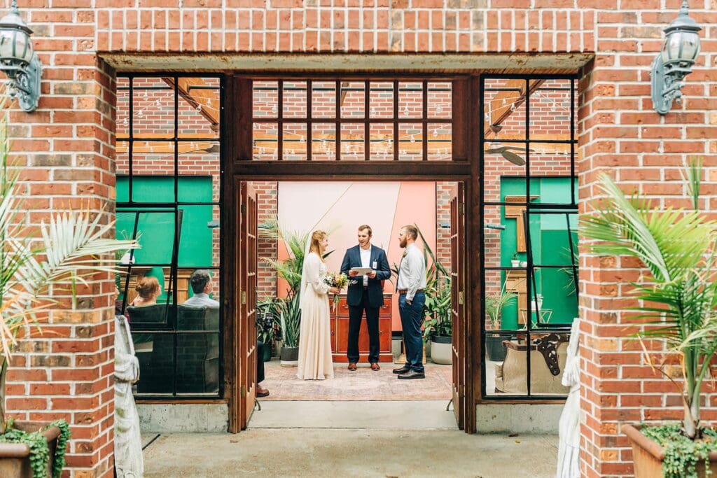 A couple is seen through a glass doorway exchanging vows in an all-brick venue space