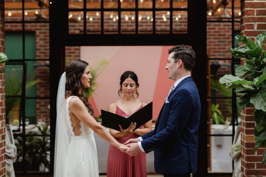 A bride and groom hold hands as they exchange vows in a courtyard venue