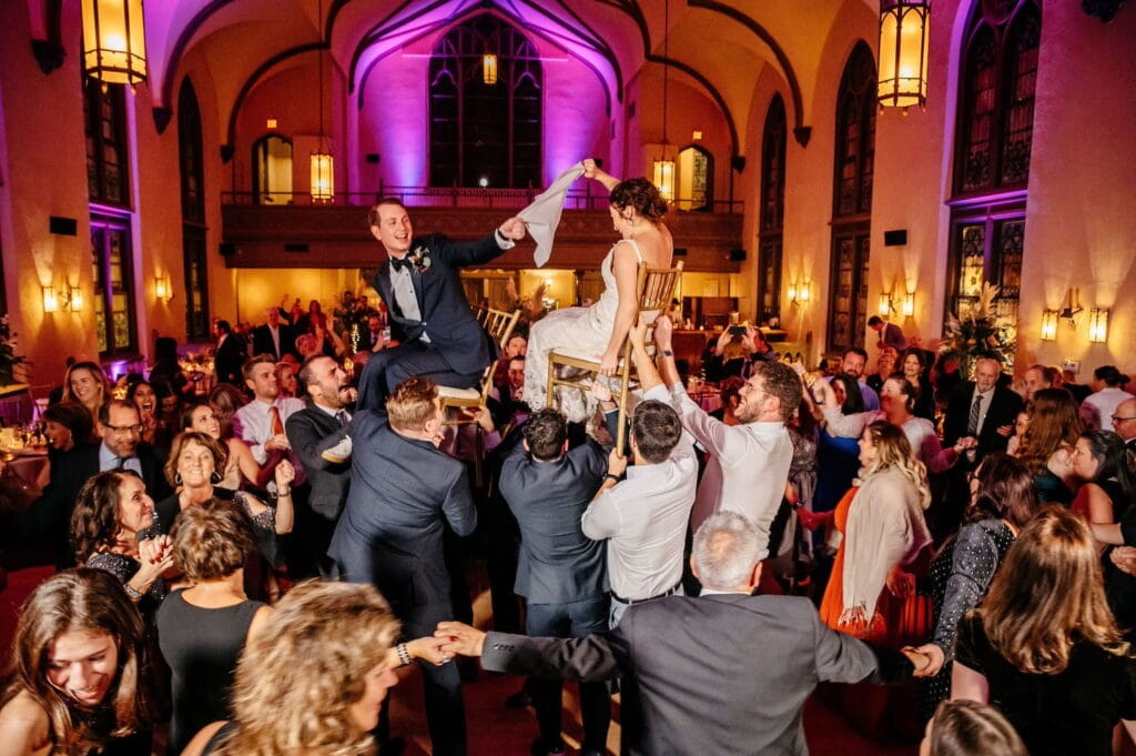 A bride and groom are raised in the air on chairs in a traditional Hora in a large reception venue