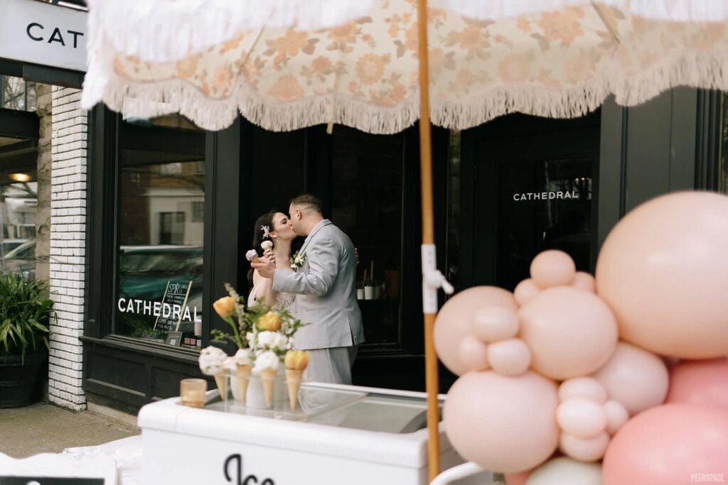 A couple kisses while holding ice cream cones standing outside on the sidewalk in front of a building