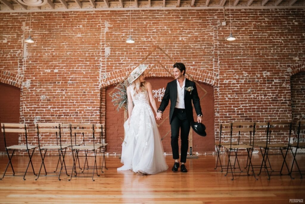 A bride and groom smile at each other as they walk hand in hand down the aisle at an industrial venue with exposed brick walls