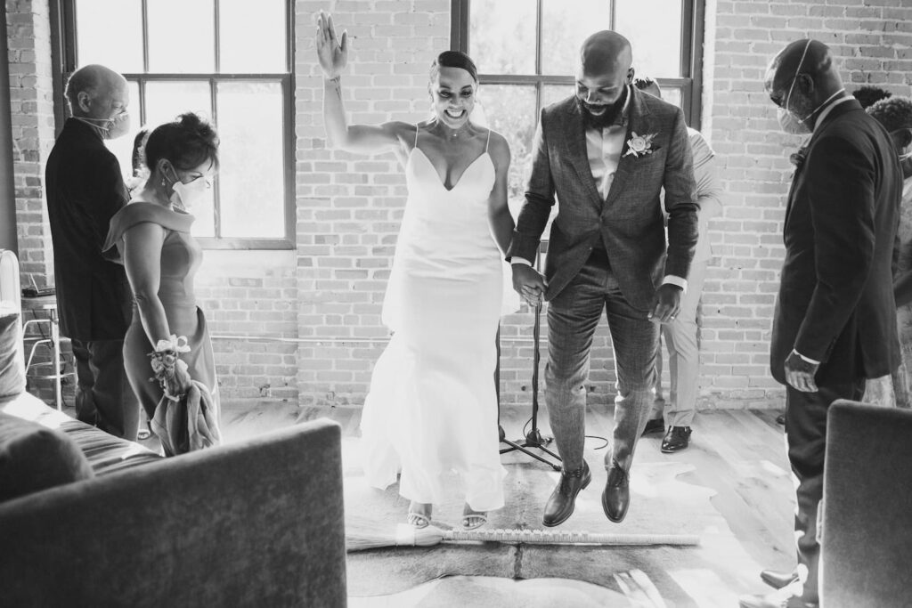 A newly-married couple jumps the broom as they make their way from the altar back down the aisle in an industrial-style wedding venue