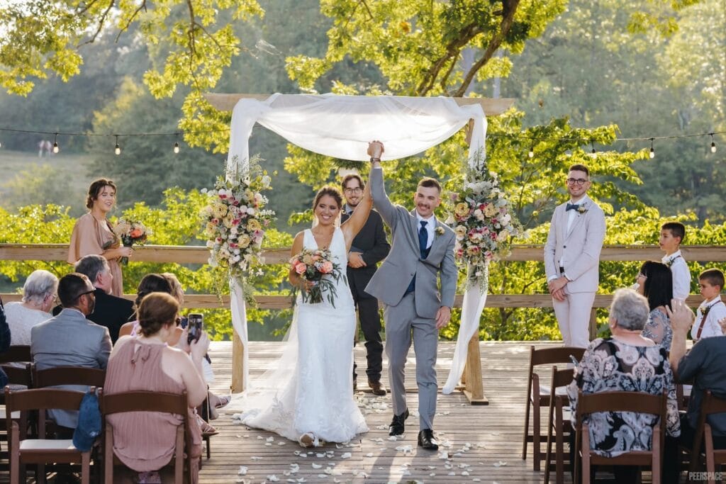 A just-wed couple walks hand in hand down an aisle full of friends and family in an outdoor venue