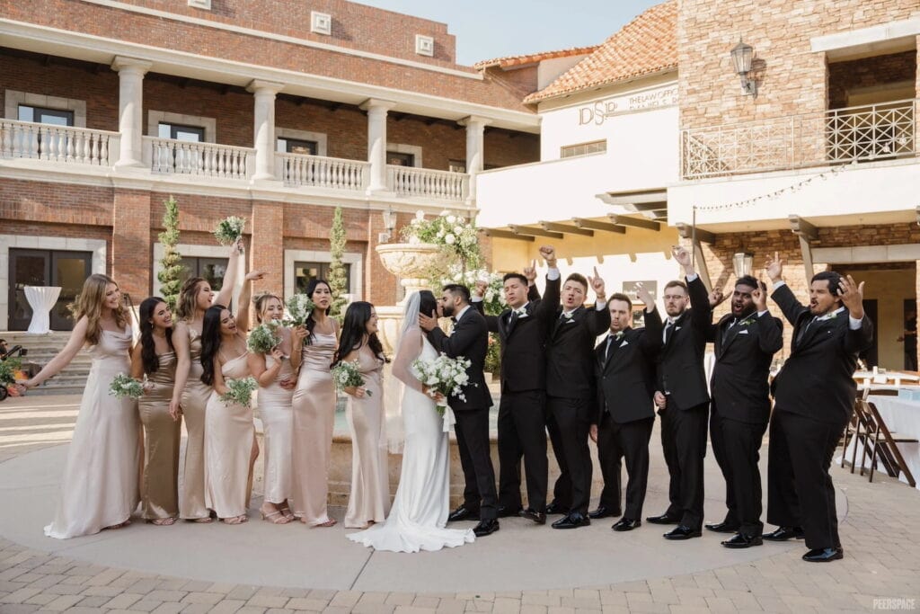 A bridal party cheers as a bride and groom kiss in the courtyard of a grand wedding venue