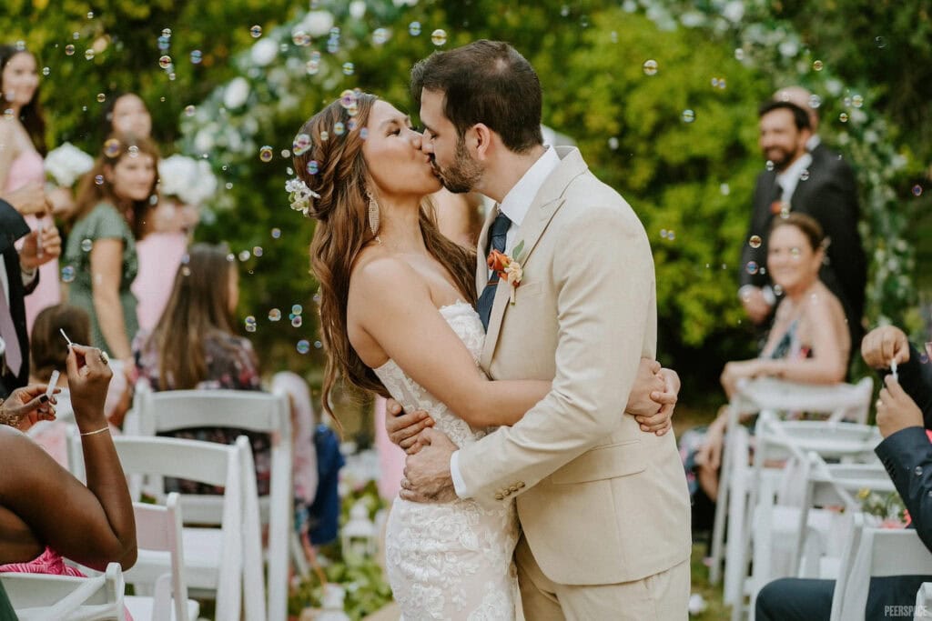A bride and groom kiss at the end of the aisle while onlookers blow bubbles, which surround them