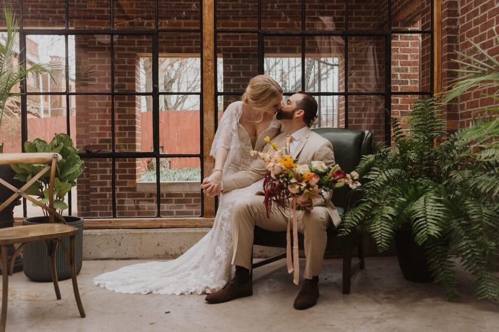 A bride in a romantic lace dress kisses her husband, dressed in tan suit, as he holds her bouquet while seated in a leather chair