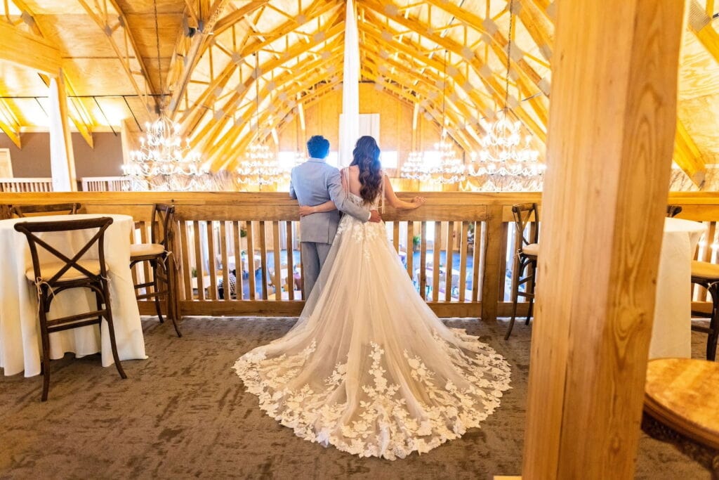 A bride and groom stand arm-in-arm on a balcony of a barn admiring their reception space below