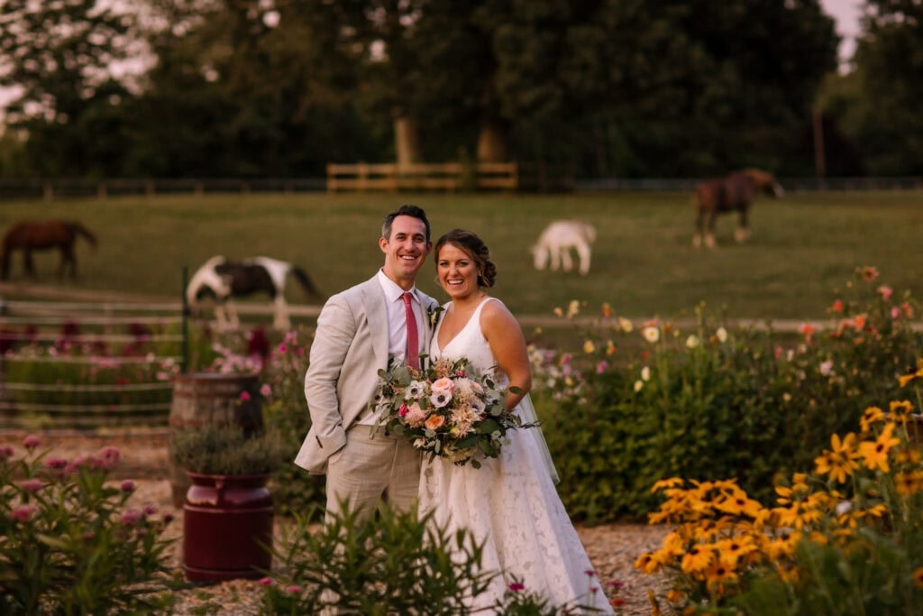 A newlywed couple pose on a farm with horses in the background