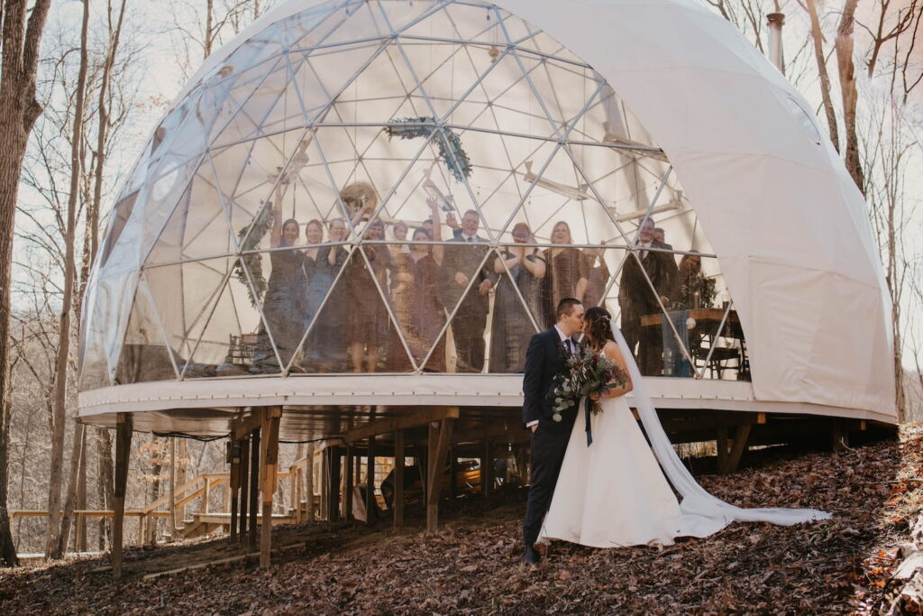 A couple shares a kiss outside of a large dome structure where friends and family look on and cheer