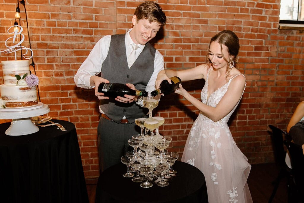 A newly-wedded bride and groom pour champagne into a tower of champagne glasses next to a cake table
