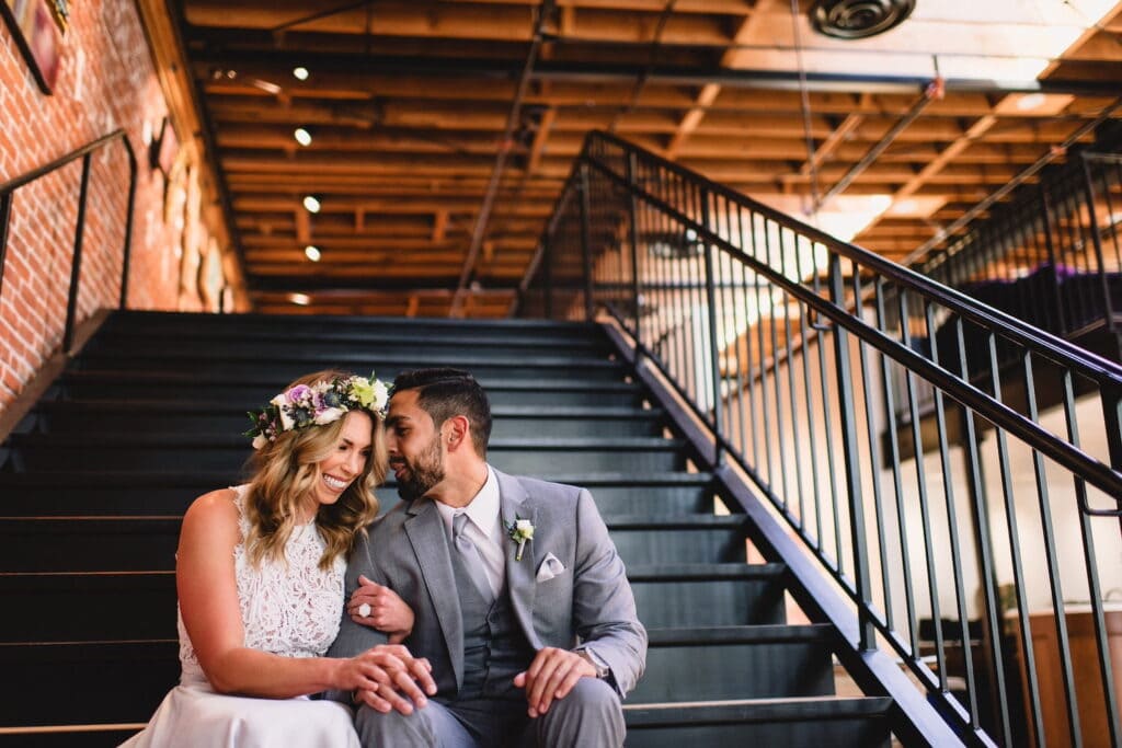 A bride in a flower crown and her groom sit on the stairs in an industrial venue and share a laugh