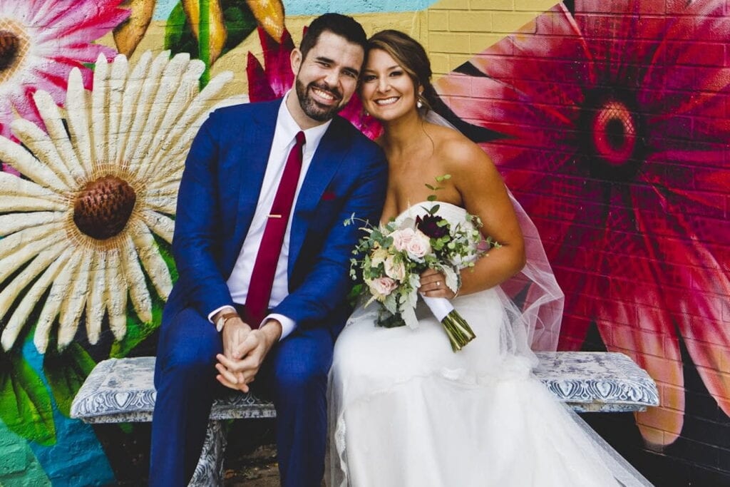 A smiling bride and groom sit on a bench in front of a large floral mural
