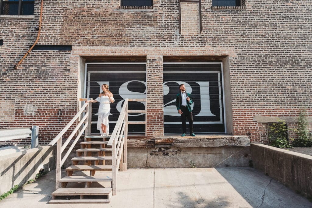 A bride and groom stand in separate truck bays in a brick building with doors painted in black and white