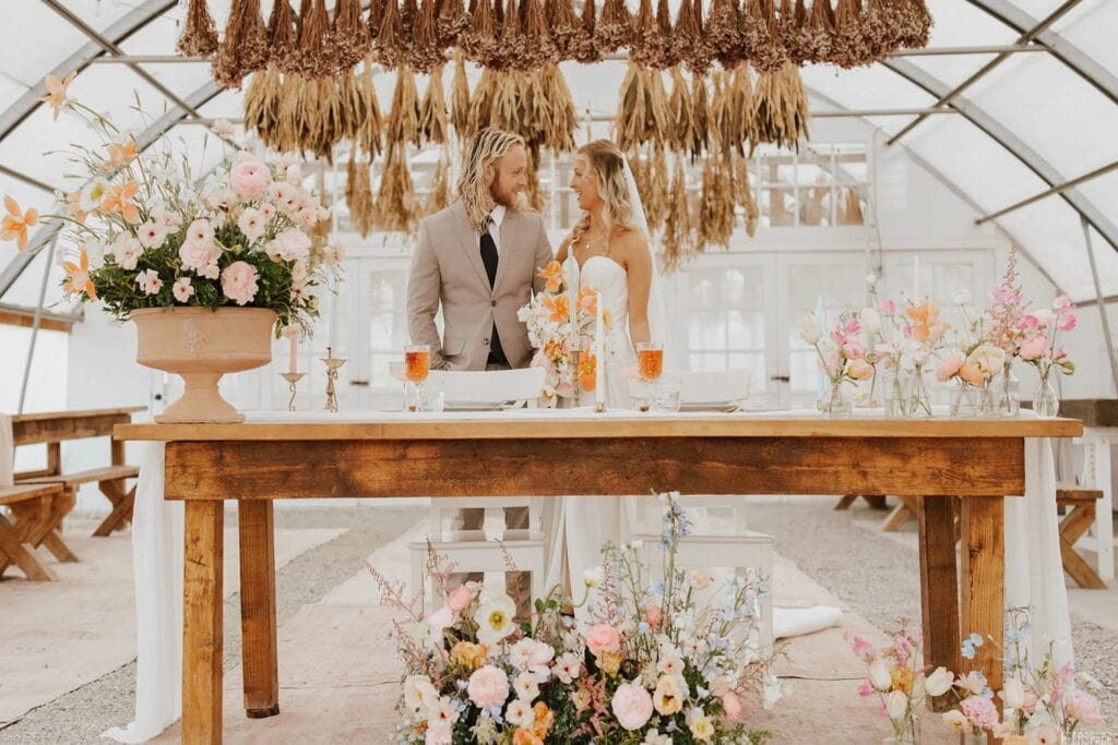 A bride and groom stand beneath dried flowers by a wooden table covered in pink and orange flowers