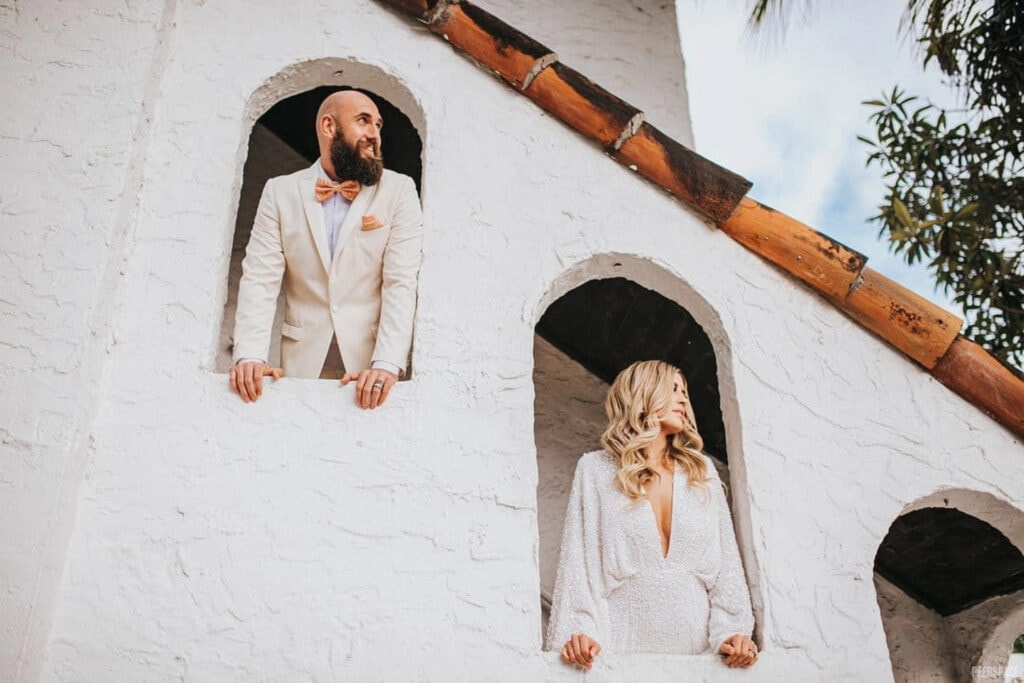 A bride and groom stand in arched windows in a Spanish style home.