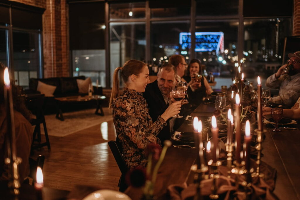 A couple toasts while sitting a dark wooden table in a loft space lit by candlelight