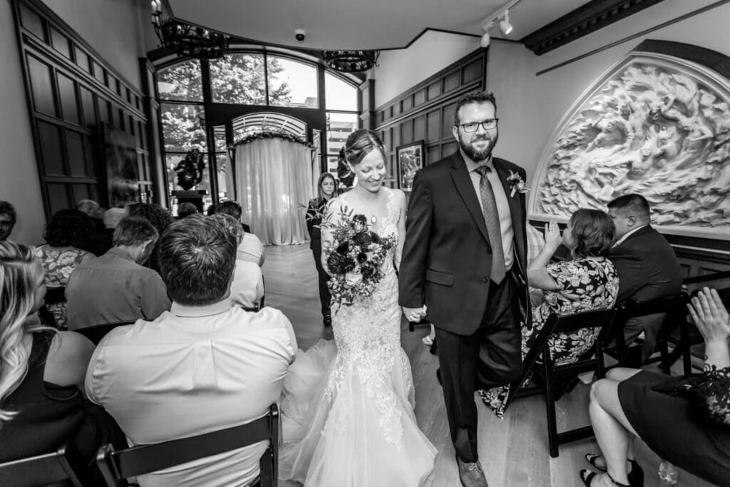 A couple walks down the aisle set up in the foyer of grand mansion as guests clap