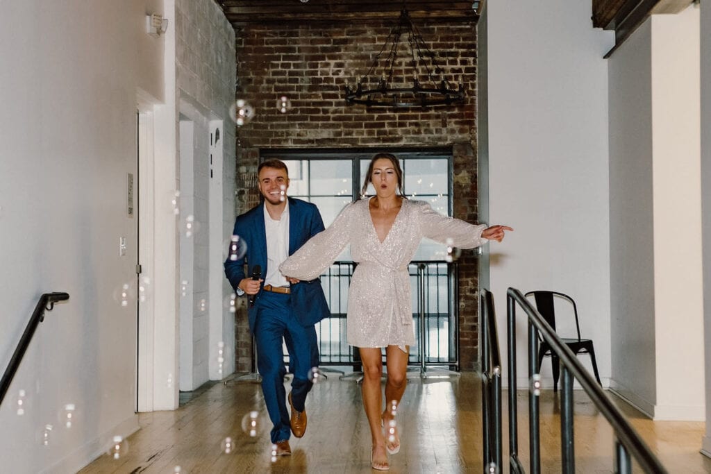 A couple joyfully walks towards a staircase in casual wedding attire with bubbles floating in the air