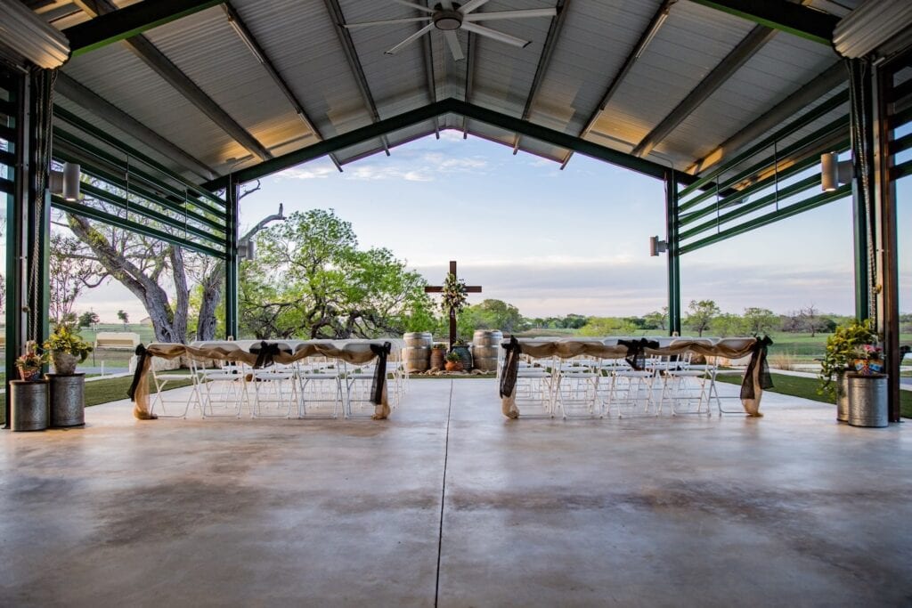 Outdoor wedding ceremony set up under metal pergola facing open field with large cross at altar set among wine barrels
