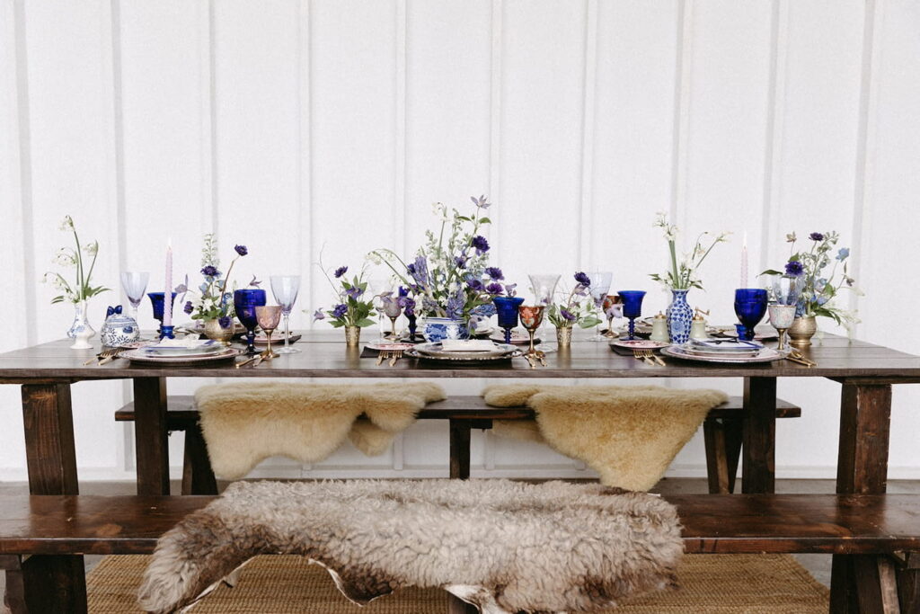 A dark wood dining table is set with purple floral arrangements, tall candles, blue glassware and plush cushions on the benches
