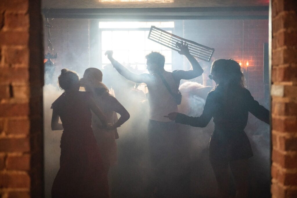 Silhouettes of people dancing in a smoky brick venue illuminated by a bright window