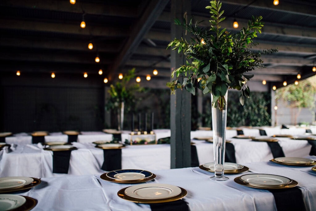 Tall glass vases with simple green florals decorate long reception tables covered in white linens in a dark, industrial reception space open to the outdoors
