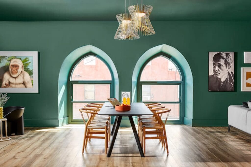 Green walls set the tone for a moody dining space punctuated by two arched windows and unique pieces of art