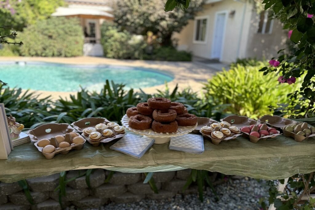 A dessert table is set up on a table outside by a pool glistening in the sunshine