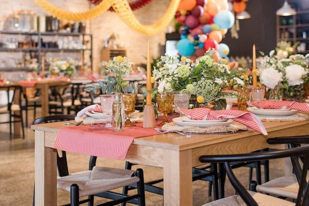 Closeup of a table set for a dinner party with red and pink striped linens and lush table arrangements with a colorful balloon arch in the background