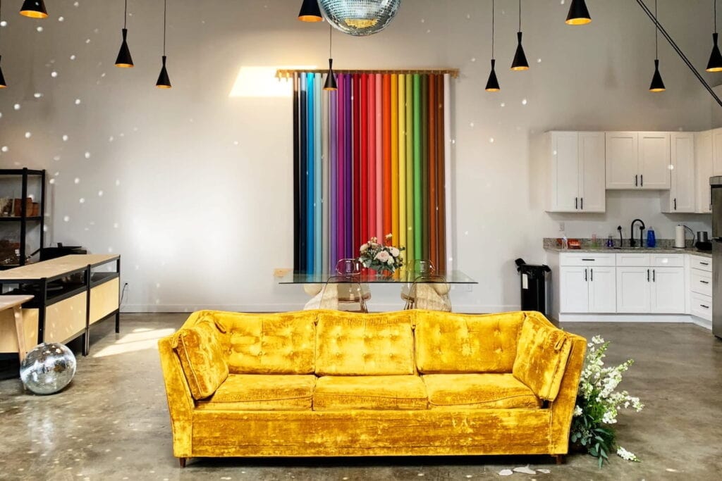 A yellow velvet couch sits in the middle of a photo studio with a kitchenette and a rack of different colored background paper hanging on the wall